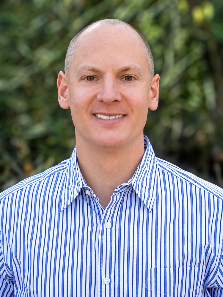 Headshot of a smiling man wearing a blue-and-white striped shirt outdoors.