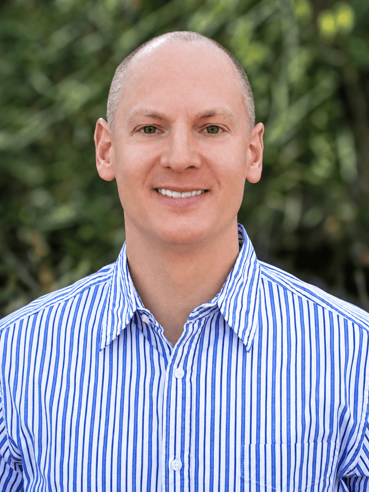 Professional headshot of a smiling man in a blue-and-white striped shirt with blurred green foliage in the background.