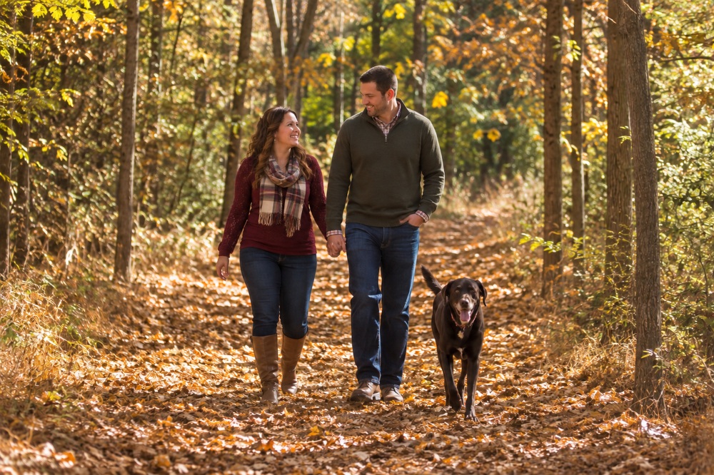Couple walking their dog on a leaf-covered trail in a wooded area during fall.